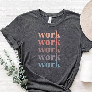 Op de afbeelding: Een donker antracietgrijs t-shirt met korte mouwen waarop het woord work vijf keer in een verticale kolom is gedrukt, waarbij elke regel een kleurverloop heeft in koraal, mauve, lavendel en lichtblauw.