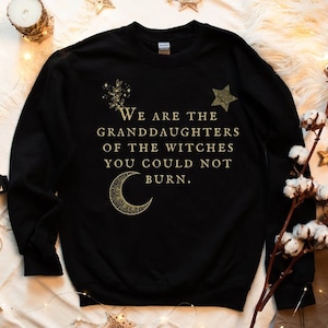 Op de afbeelding: Zwarte sweatshirt met gouden tekst "We are the granddaughters of the witches you could not burn". Het ontwerp bevat een maan, een ster en bloemmotieven. De sweatshirt ligt op een witte, zachte ondergrond.