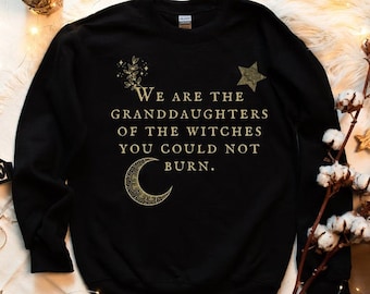 Może przedstawiać: Czarna bluza z napisem "We are the granddaughters of the witches you could not burn" w złotym kolorze. Projekt zawiera półksiężyc, gwiazdę i elementy kwiatowe. Bluza leży na białej, puszystej powierzchni.