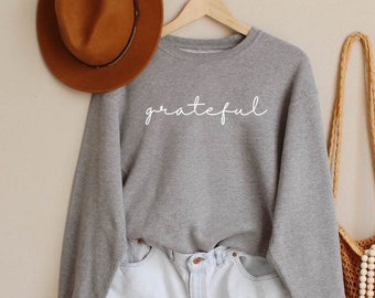 May include: A grey crewneck sweatshirt with the word grateful printed in white script lettering, paired with light blue denim shorts, a brown felt fedora hat, and a tan crocheted tote bag with a long wooden bead tassel accessory.