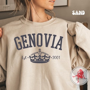 Op de afbeelding: Zandkleurige sweatshirt met het woord "GENOVIA" in marineblauwe blokletters. Daaronder een marineblauw kroonontwerp met de tekst "Est. 2001". De sweatshirt heeft een ronde hals.