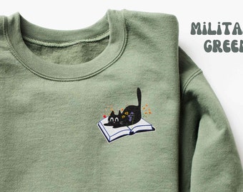 May include: A military green crewneck sweatshirt featuring an embroidered graphic of a black cat lying on an open book, surrounded by small colorful flowers. The text "MILITARY GREEN" is printed in a rounded, bold font in the upper right corner.