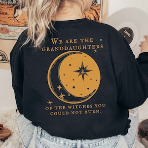 Op de afbeelding: Zwart sweatshirt met gouden tekst en afbeelding. De achterkant van de sweatshirt heeft de tekst "We are the granddaughters of the witches you could not burn". Een gouden halve maan en sterrenafbeelding is gecentreerd onder de tekst.