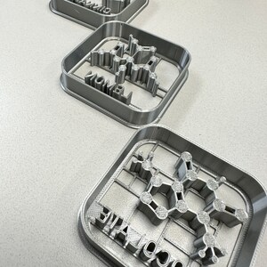 May include: Three gray cookie cutters in the shape of molecular structures. Each cutter has a different word: "Cinnamon", "Vanilla", and "Chocolate". The cutters are square with a grid pattern.