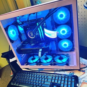 May include: A pink computer case with a clear side panel, showcasing a custom-built computer with blue LED lights and multiple fans. The case is sitting on a desk with a black keyboard.