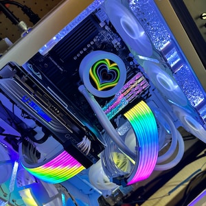 May include: A white computer case with a clear side panel, revealing a rainbow-colored cable management system. The motherboard has a heart-shaped light and a white fan with a green, yellow, and blue heart design.