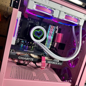 May include: Pink computer case with a clear panel showing the interior components, including a graphics card with the text "GIGABYTE GEFORCE RTX" and a motherboard with a heart-shaped LED light. The case has a mesh panel on the bottom and a white fan with pink accents.