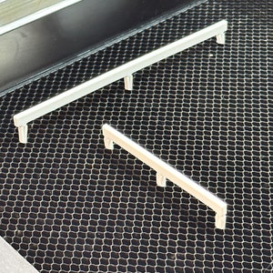 May include: Two silver metal bars with rounded ends, resting on a black wire mesh surface. The bars are positioned at an angle to each other.