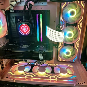 May include: A pink computer case with a clear window showing the interior. The case has a black graphics card with a rainbow-colored fan and a heart-shaped light. The case also has a black motherboard with a white cable. The case is sitting on a black keyboard.