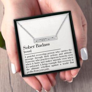 May include: A silver-toned necklace with a rectangular pendant featuring Morse code and the words "Sober Badass." The necklace is presented in a black gift box with a white interior, and the definition of "Sober Badass" is printed on the card.