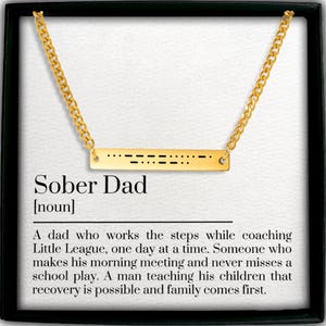 May include: Gold-colored necklace with a bar pendant featuring Morse code and the words "Sober Dad." The necklace is displayed in a black framed box with text describing a sober father.