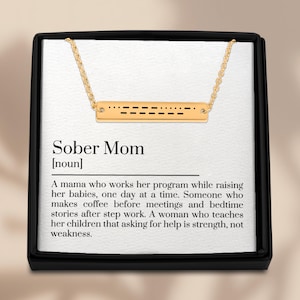 May include: A gold-colored necklace with a rectangular pendant featuring a Morse code design. The necklace is presented in a black box with the words "Sober Mom" and a definition printed on the inside.