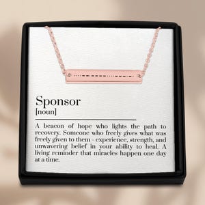 Op de afbeelding: Een roségouden staafketting met een fijne ketting, gepresenteerd in een zwarte geschenkdoos. De staaf heeft een patroon van stippen en streepjes. De doos bevat de definitie van "Sponsor".