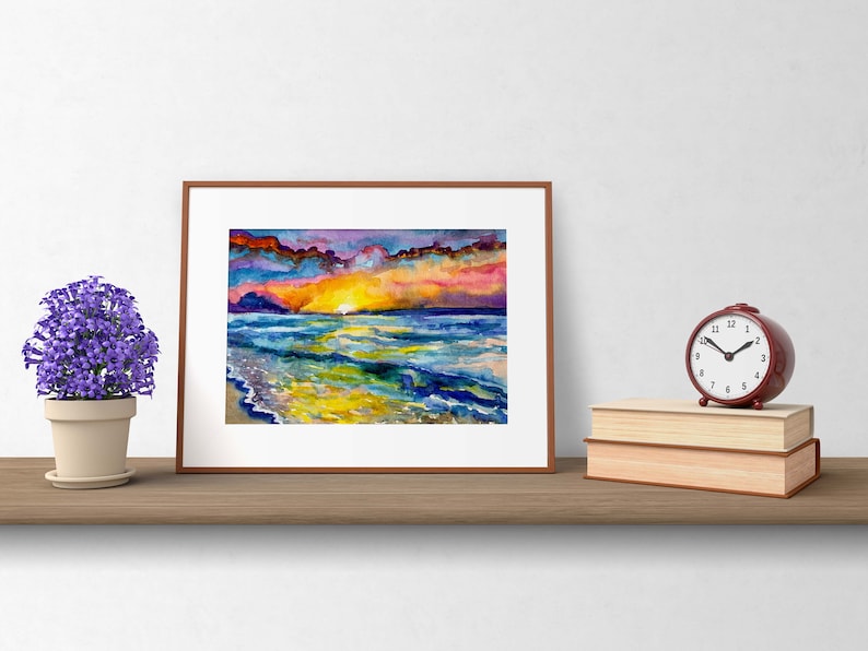 Op de afbeelding: Een ingelijst aquarel schilderij van een zonsondergang boven de oceaan. De lucht is een mix van paars, roze en oranje, met wolken die de kleuren reflecteren. De oceaan is diepblauw, met golven die breken op de kust. Het zand is lichtgeel.