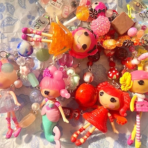 May include: A selection of colourful doll charms featuring mermaid and dress-wearing figures. The charms are embellished with beads and other decorative elements, creating a playful and whimsical aesthetic.
