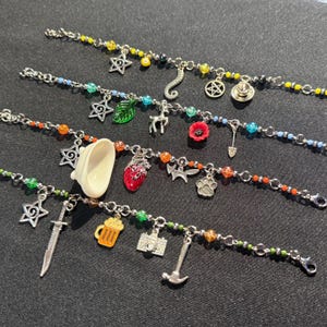 May include: Four silver-tone charm bracelets featuring colorful beads and various metal pendants, including stars, a heart, a sword, a camera, a hammer, a beer mug, a paw print, a leaf, a flower, a pentagram, and a small white ceramic bathtub.