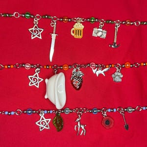 May include: Three silver charm bracelets with various charms, including stars, a sword, a beer mug, a camera, a hammer, a heart, a bird, a paw print, a leaf, a deer, a poppy, and a shovel. The bracelets have colorful beads and are set against a red background.