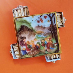 May include: A square decorative box with a gold-toned frame featuring a colorful illustration of figures in a forest setting with autumn leaves. The box is surrounded by four open matchboxes filled with wooden matches, each with a small wooden bead handle.