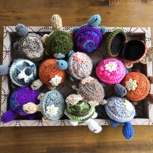 May include: A collection of crocheted turtle figurines in a variety of colors, including purple, pink, green, orange, and brown. The turtles are arranged in a wooden box with a patterned lining.