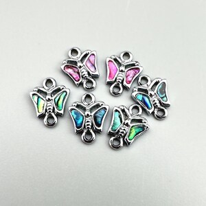 May include: Six silver-toned metal butterfly charms featuring iridescent shell inlays in shades of pink, blue, and green. Each charm has a symmetrical butterfly shape with a small circular loop at the top and bottom for attachment to jewelry.