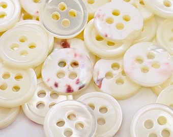 May include: A collection of round, four-hole buttons in shades of off-white and cream. Some buttons feature a smooth, solid finish, while others display subtle, mottled pink and brown patterns with a pearlescent, iridescent sheen.