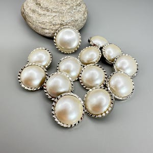 May include: A collection of pearl-like buttons with silver-colored, textured rims. The buttons are round and have a shiny, off-white appearance. A large, textured, gray stone is in the background. The buttons are scattered on a gray surface.