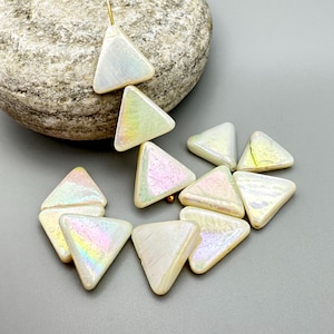 May include: Iridescent triangular beads in various sizes. The beads have a pearlescent finish that reflects multiple colors. Some beads are strung on a gold-colored wire. The beads are arranged on a gray surface.