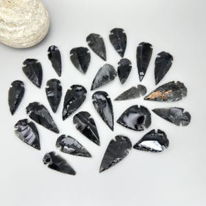 May include: A collection of obsidian arrowheads in various shapes and sizes, displayed on a white surface. The arrowheads are primarily black with some showing gray and brown inclusions. A large, rounded, light-colored stone is in the upper left corner.
