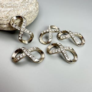 May include: Five silver-toned infinity charms, each encrusted with clear rhinestones. The charms are arranged on a light gray surface, with a large, textured rock in the background. The infinity symbol is a popular design element.