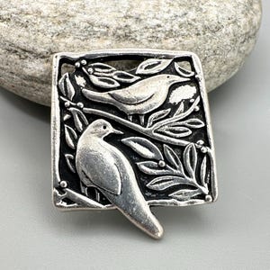 Peut inclure: Broche carrée argentée représentant deux oiseaux perchés sur des branches feuillues. Les oiseaux sont détaillés avec des plumes et des becs visibles. La broche a une bordure sombre et est posée sur un fond neutre.