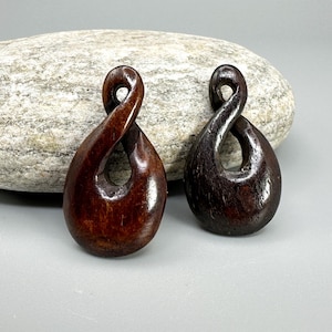 May include: Two wooden pendants with a teardrop shape and an infinity symbol design. The pendants are a dark brown color and have a smooth, polished finish. The background is a gray surface with a large rock.