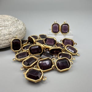 May include: A collection of dark purple, rectangular gemstone pendants with gold-colored metal frames. Two matching earrings are displayed upright. The gemstones have a faceted appearance. The pendants have small loops for attaching to jewelry.