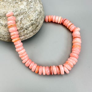 May include: A strand of pink and peach-colored beads, likely for jewelry making, is displayed against a gray background. The beads are disc-shaped and vary slightly in color, creating a gradient effect. A large, textured rock sits behind the beads.