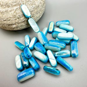 May include: A collection of blue and white oval-shaped beads. The beads have a glossy finish and are strung together on a white string. The beads are scattered on a gray surface, with a large rock in the background.