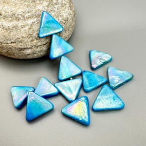 May include: A collection of iridescent blue triangle beads. The beads have a glossy finish and a range of blue hues, with hints of yellow and pink. The beads are scattered on a gray surface.