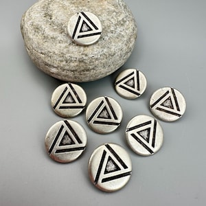 May include: A collection of silver-toned metal buttons, each featuring a black triangle design within a circle. The buttons are scattered on a gray surface, with one resting on a textured gray stone. The buttons have a vintage or antique appearance.