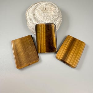 May include: Three rectangular tiger's eye stones with a polished finish, displaying bands of brown and gold. A rounded, gray stone sits behind the rectangular stones. The stones are arranged on a light gray surface.