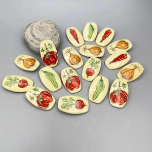 May include: A selection of small, oblong ceramic dishes featuring hand-painted vegetable designs. The vegetables include tomatoes, onions, peppers, and radishes, in vibrant colours against a cream background. A grey stone sits in the background.