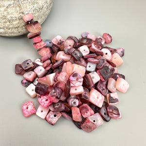 May include: A collection of pink, white, and dark red square-shaped beads with holes. Some beads are strung on a white string. The beads are scattered on a light grey surface. The beads are made of a shell-like material.