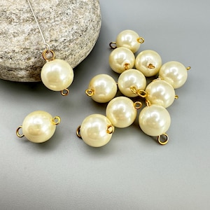 May include: Ivory-colored faux pearl beads with gold-tone loops. The beads are round and have a glossy finish. One bead is attached to a silver-tone pin. The beads are scattered on a gray surface, with a large rock in the background.