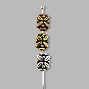 May include: Three decorative beads in copper, gold, and silver tones are strung on a silver pin. Each bead features a symmetrical floral design with raised details. The beads are displayed against a gray background.