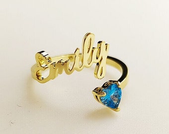 Op de afbeelding: Gouden ring met de naam "Emily" in cursief en een blauwe hartvormige edelsteen.