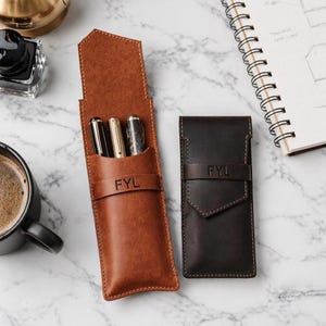 May include: Two leather pen cases, one tan and one dark brown, each featuring a flap closure and a horizontal strap embossed with the letters FYL. The tan case is open, revealing three pens inside, while the dark brown case is closed.