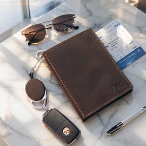 May include: A brown leather wallet with the initials "FYL" embossed on the front, a pair of sunglasses, a car key, a pen, and a boarding pass. The wallet is rectangular with stitched edges. The sunglasses have a gold frame and brown lenses.