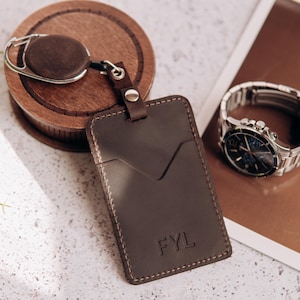 May include: A dark brown leather badge holder with a V-shaped pocket and the embossed letters FYL. The holder is attached to a retractable reel with a silver metal clip. A silver-tone wristwatch with a dark face is visible nearby.