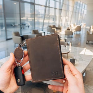May include: A brown leather wallet with the initials "FYL" embossed on the front. The wallet is held next to a car key with a black remote and a brown key fob. The wallet has a stitched border.