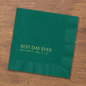 May include: A square dark green paper napkin featuring gold foil text that reads BEST DAY EVER followed by the names MAX & MYLA and the date 09 | 12 | 25. The napkin has a textured border along the right edge and a smooth center surface.