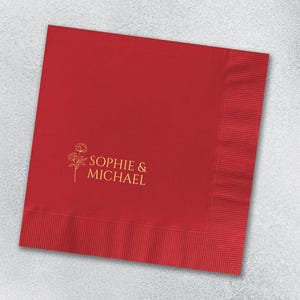 May include: A square red paper napkin featuring a gold-colored line art illustration of two flowers and the text "SOPHIE & MICHAEL" printed in a clean, sans-serif font. The napkin has a textured, embossed border along the right and bottom edges.