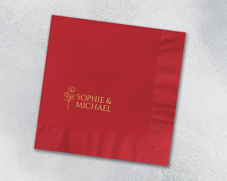 May include: A square red paper napkin featuring a gold-colored line art illustration of two flowers and the text "SOPHIE & MICHAEL" printed in a clean, sans-serif font. The napkin has a textured, embossed border along the right and bottom edges.