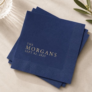 May include: A stack of square navy blue paper napkins with a textured border. The top napkin features gold foil text that reads THE MORGANS and SEPT 02, 2025, centered in a serif font. The napkins are layered slightly offset from one another.